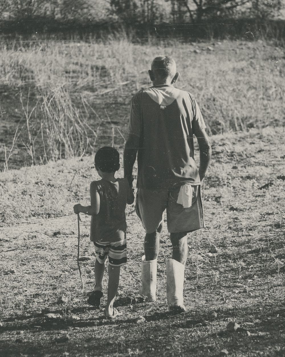A father and his young son walking hand in hand across a field, in black and white